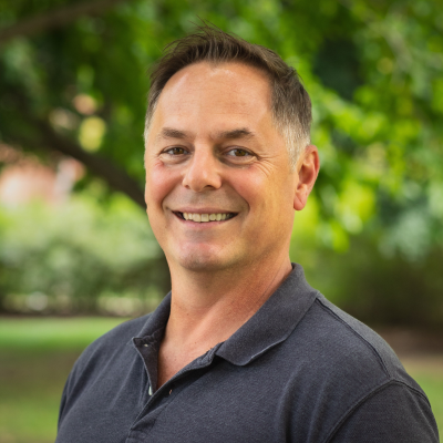 Portrait of Eric Benbow outdoors, wearing a dark short‑sleeve collared shirt, standing in front of leafy green trees with a softly blurred background